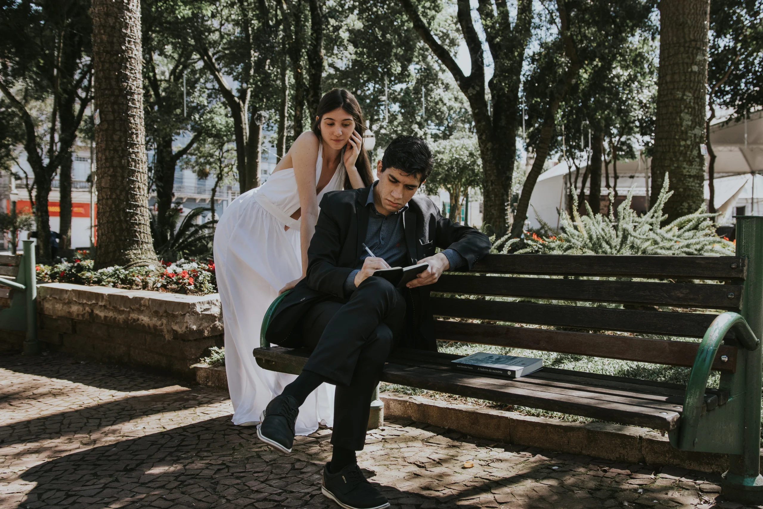 O autor sentado num banco de praça, escrevendo, e Mariana escorada atrás dele, contemplando.